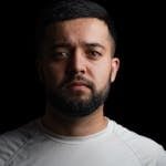 Studio portrait of a serious man with a beard in a white t-shirt against a black backdrop.